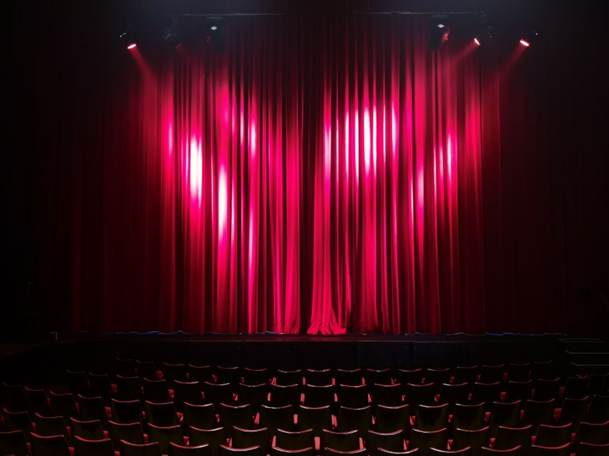 A spacious auditorium with multiple tiers of curved wooden balconies and orange upholstered seats, viewed from the stage area. The seating is arranged in a semi-circular pattern, with several rows on 